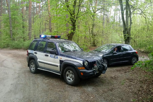 W lesie na skrzyżowaniu dróg stoi stoi policyjny radiowóz, a obok ciemny samochód, w którym siedzi mężczyzna.