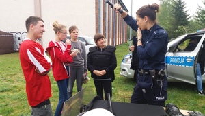 Zewnętrzny teren szkoły. Policjantka rozmawia z uczestnikami konkursu pokazując sprzęt będący na wyposażeniu funkcjonariusza.
