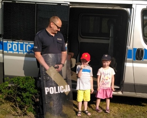 Policjant prezentuje dzieciom sprzęt policyjny. Tło stanowi radiowóz.