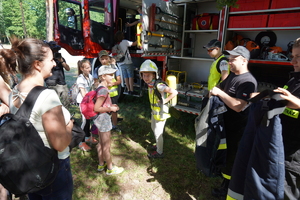 Dzieci przymierzają elementy munduru strażackiego. Tłem wóz strażacki bojowy.