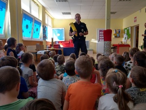 Policjant rozmawia z dziećmi w przedszkolu o bezpiecznym wypoczynku trzymając bojkę ratownicza.. Dzieci siedzą na podłodze.