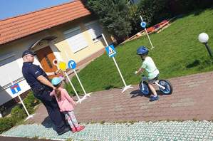 Dzieci pod okiem policjanta ćwiczą jazdę rowerkami na terenie przedszkola, gdzie ustawione są znaki drogowe.