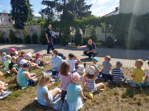 Policjanci rozmawiają z dziećmi siedzącymi na trawie. Jeden z policjantów kuca przed dziećmi.