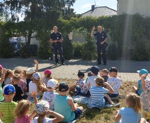 Policjanci stoją przed dziećmi siedzącymi na trawie i z nimi rozmawiają.