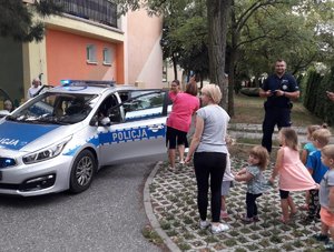 Policjant prezentuje dzieciom radiowóz policyjny zaparkowany przed placówką.