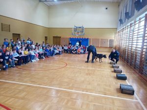 Policjant prezentuje umiejętności psa służbowego podczas prelekcji na sali gimnastycznej.
