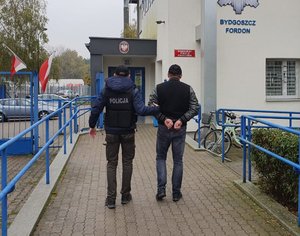 Policjant prowadzi zatrzymanego. W tle wejście do Komisariatu  Policji Bydgoszcz- Fordon.