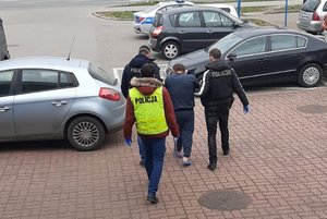 trzej policjanci prowadzą zatrzymanego skutego kajdankami zespolonymi