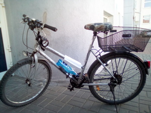 rower oparty na nóżce stoi na chodniku. Na ramie ma włożoną w koszyk od bidonu butelkę z wodą
