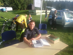 Policjant podczas działań w Myślęcinku. Siedzi przy stoliku z dokumentami do znakowania rowerów. W tle rowerzyści.