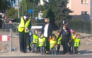 Dzieci wraz z opiekunkami przechodzą przez ulicę pod okiem policjantki idącej z nimi.