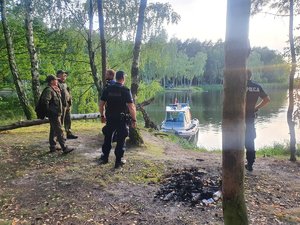 Policjanci w lesie przy miejscu palenia się ogniska. W tle policyjna łódź motorowa na wodzie.