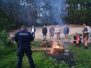 Policjant stoi przy grupie osób, które rozpaliły ognisko w lesie.