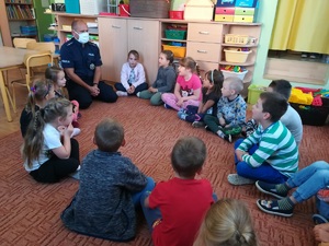 Policjant wraz z dziećmi siedzą w okręgu na dywanie w sali przedszkolnej.