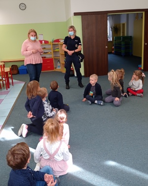 Policjantka i wychowawczyni prowadzą zajęcia z dziećmi siedzącymi na dywanie w sali przedszkolnej.