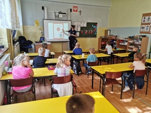 Policjantka prowadzi zajęcia z uczniami w klasie szkolnej. Policjantka pokazuje w stronę wyświetlanego na tablicy slajdu.