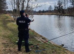Policjant stoi przy wędkarzu, który łowi ryby w rzece.
