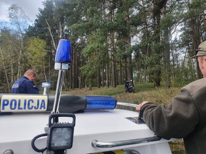 Policjant i leśnik na policyjnej łodzi motorowej. W tle las i zaparkowany w nim motocykl.