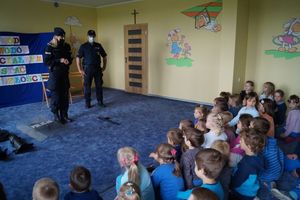 Policjanci stojąc przed dziećmi siedzącymi na dywanie w sali przedszkola prowadzą prelekcję.