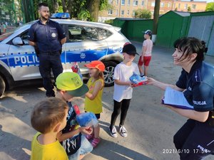policjantka rozdaje dzieciom maskotki Polfinki. W tle stoi policjant koło radiowozu