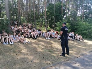 Policjant stojąc przed uczniami siedzącymi na trawie prowadzi prelekcję. W tle las.