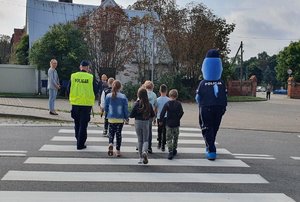 Dzieci wraz z policyjnym Polfinkiem przechodzą przez przejście dla pieszych.
