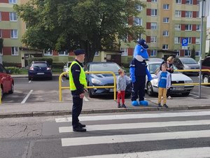 Policjant stoi na pasach, a Polfinek z dwoma chłopcami stoi przy przejściu.