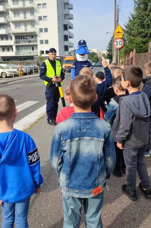 Policjant wraz z Polfinkiem rozmawiają z uczniami przy przejściu dla pieszych.