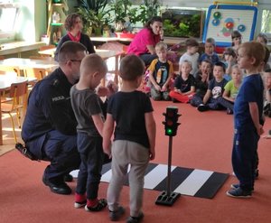 Policjant prowadzi zajęcia z dziećmi używając maty imitującej przejście dla pieszych z sygnalizatorem. Troje dzieci stoi przy nim.