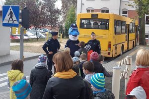 Policjanci wraz z Polfinkiem rozmawiają z uczniami  na terenie przyszkolnym. W tle autobus szkolny.
