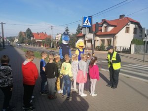 Dzieci wraz z policjantami stoją przed przejściem dla pieszych. Policjantka rozmawia z dziećmi.
