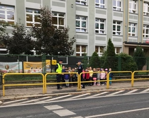 Dzieci wraz z policjantką idą chodnikiem w stronę przejścia dla pieszych.
