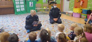 Policjantka czyta dzieciom książkę. Obok siedzi drugi policjant. Przed nimi siedzą w półkolu dzieci.