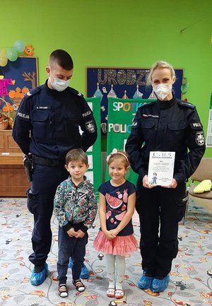 Policjantka i policjant oraz dwoje dzieci stoją i pozują do zdjęcia w sali przedszkolnej. Policjantka trzyma laurkę z podziękowaniami.