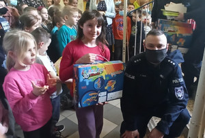 Policjantka kuca przy dziewczynce trzymającej nagrodę. W tle stoją inne dzieci.
