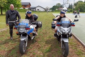 dwaj chłopcy siedzą na dwóch policyjnych motocyklach. Obok stoi policjant