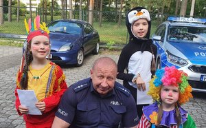 Policjant kuca, a przy nim stoją dzieci przebrane w kolorowe stroje i peruki.